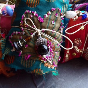 May include: Close-up of a teal fabric bundle with a heart-shaped embellishment. The heart is outlined with pink sequins and embroidery. A white string is tied around the bundle, with a black bead and silver charm hanging from it. The bundle is part of a collection.