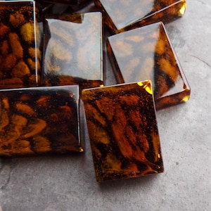 May include: A group of eight rectangular, translucent, amber-colored glass tiles with a brown marbled pattern. The tiles are arranged in a random pattern on a gray surface.