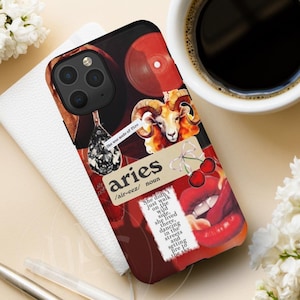 May include: A phone case with a collage design featuring a ram, red lips, cherries, and the word "aries". The case has a black border and a dark red background. Text on the case reads "She was made of fire" and "She didn't just walk on the wild side, she lived there..."