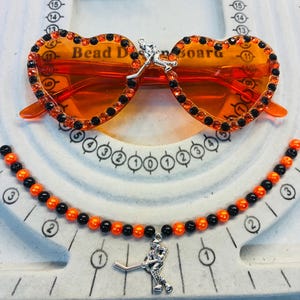 May include: Orange heart-shaped sunglasses with black and orange rhinestones. A matching necklace features orange and black beads and a hockey player charm. The sunglasses have the text "Bead Denora".