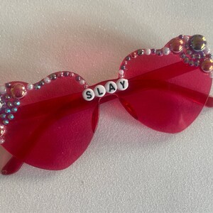 May include: Pink heart-shaped sunglasses with rhinestones and the word "SLAY" spelled out in white letters.
