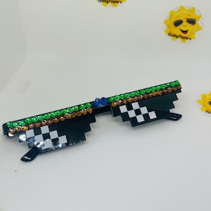 May include: Black plastic sunglasses with a chequered pattern on the lenses. The sunglasses are decorated with green, orange, and blue rhinestones.