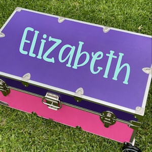 May include: A purple and pink storage trunk with silver hardware and the name "Elizabeth" written in turquoise lettering on the lid.