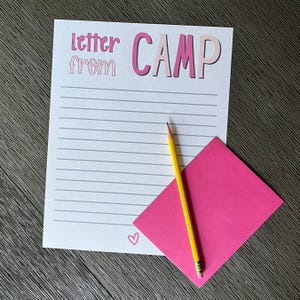 May include: A white stationery sheet with horizontal gray lines, featuring the pink text "letter from CAMP" at the top and a small pink heart at the bottom. A bright yellow pencil rests diagonally across a vibrant pink envelope on the paper.
