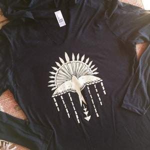 May include: Black long-sleeved shirt with a white graphic of a bird flying through a sunburst with a geometric design. The shirt has a hood and a v-neck.