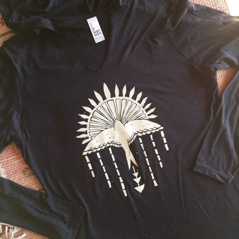 May include: Black long-sleeved shirt with a white graphic of a bird flying through a sunburst with a geometric design. The shirt has a hood and a v-neck.