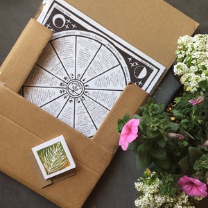Puede incluir: Una caja de cart&oacute;n marr&oacute;n con un calendario de jardiner&iacute;a blanco y negro en su interior. El calendario es una rueda con los meses y las tareas de jardiner&iacute;a enumeradas alrededor del borde. La caja est&aacute; abierta y hay dos plantas en maceta con flores blancas y rosas en la mesa junto a ella.
