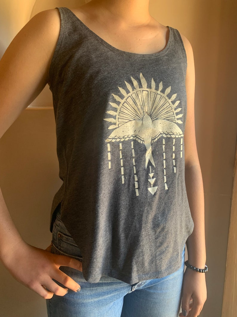 May include: A gray tank top with a white graphic of a bird flying through a sunburst design. The graphic also includes a series of vertical lines and arrows.