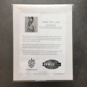 May include: A black and white photo of a woman with long dark hair wearing a red tank top and a necklace. The photo is on a white background with text that reads "Tree of Life Studio, The Artwork of Rebecca Kunz". The text below the photo describes the artist and her work. The text includes the words "Cherokee Nation", "Santa Fe", "mixed media artist", and "cultural roots". The bottom of the image includes a logo that reads "Certified New Mexico True 100% Made in NM" and the website address "treeoflifestudio.net".