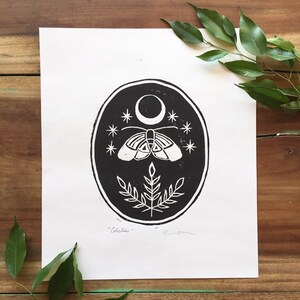 May include: Black and white print of a moth with a crescent moon and stars in a circle. The print has the word "Celestine" written below the image.