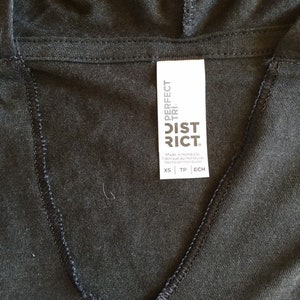 May include: A black hooded sweatshirt with a white tag that reads "PERFECT TRI. DISTRICT" and size information: XS, TP, ECH. The tag is attached to the neckline of the sweatshirt.