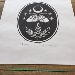 May include: Black and white print of a moth with wings spread, a crescent moon, and stars on a dark background. The print is in a circle with a plant at the bottom. The text "Celestine" is written below the circle.