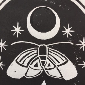 May include: Black and white linocut print of a moth with wings spread, flying under a crescent moon with stars in the background.