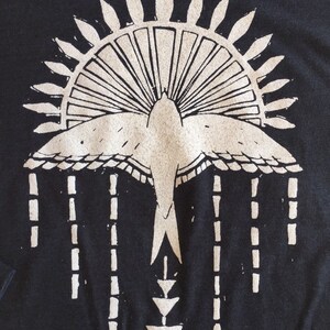 May include: A black t-shirt with a white graphic design of a stylized bird with a sunburst above it and a geometric pattern below it.