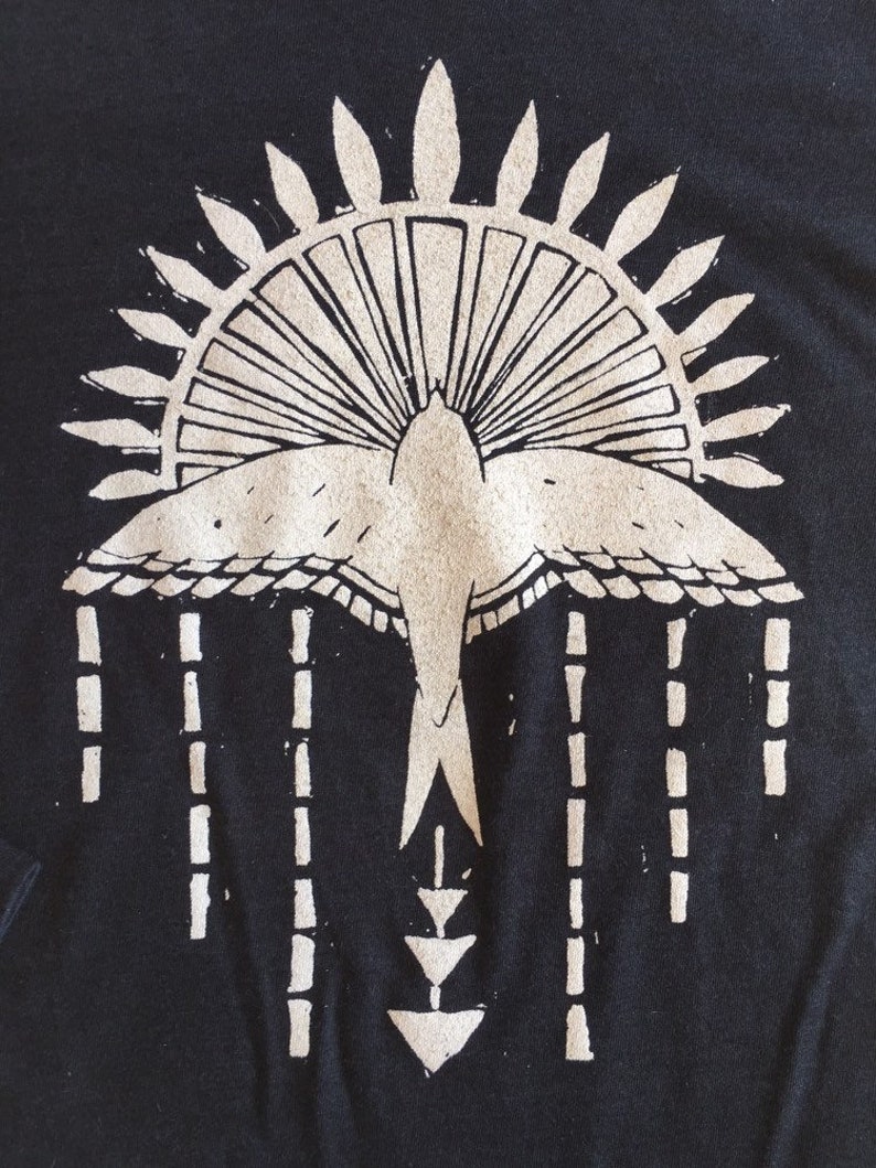May include: A black t-shirt with a white graphic design of a stylized bird with a sunburst above it and a geometric pattern below it.