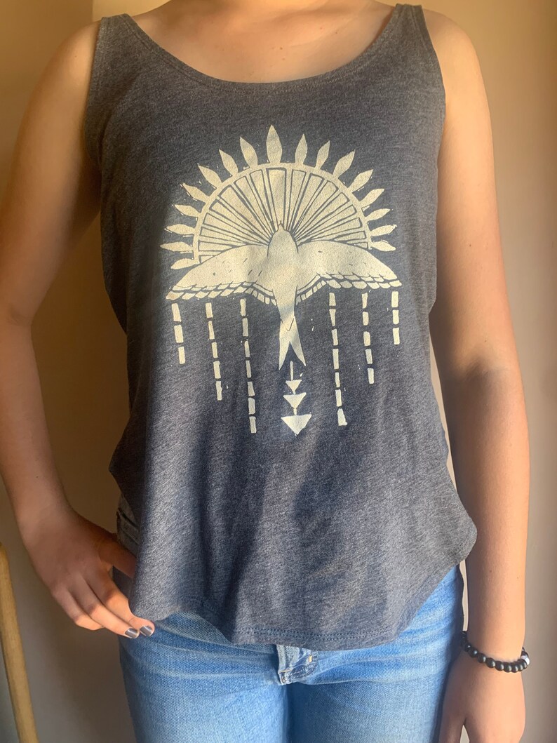 May include: A gray tank top with a white graphic of a bird flying above a sunburst design. The graphic also includes a series of vertical lines and a triangle shape.