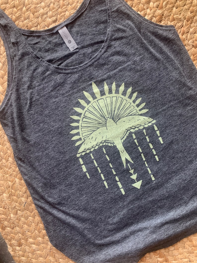 May include: A gray tank top with a green graphic of a bird flying out of a sun with a geometric design underneath.