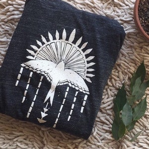 May include: A dark gray t-shirt with a white graphic print of a bird flying through a sunburst. The bird is stylized with a long tail and wings. The sunburst is made up of radiating lines.