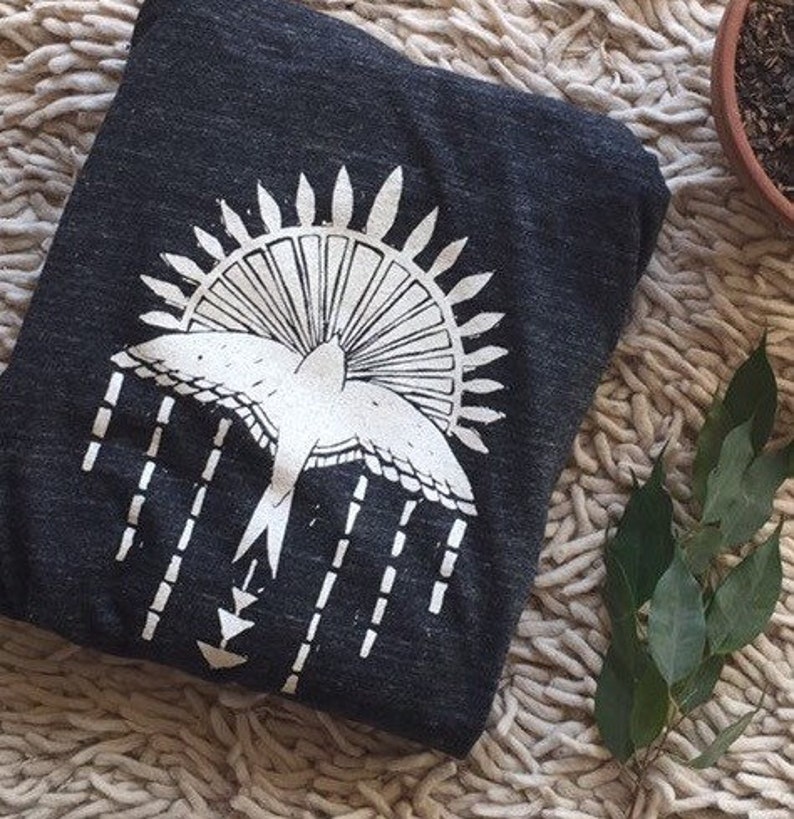 May include: A dark gray t-shirt with a white graphic print of a bird flying through a sunburst. The bird is stylized with a long tail and wings. The sunburst is made up of radiating lines.
