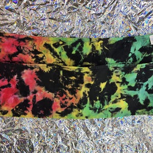 May include: A tie-dye apron with two large pockets. The apron features a vibrant mix of colors, including red, yellow, green, and black, creating a psychedelic pattern. The apron has long ties for securing around the waist.