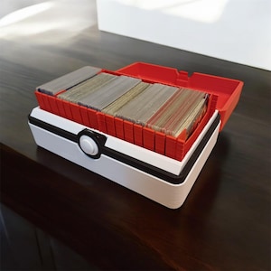 Peut inclure: Une boîte de rangement pour cartes en plastique blanche et rouge avec un motif de Poké Ball sur le devant. Le couvercle est ouvert et la boîte est remplie de cartes à collectionner.