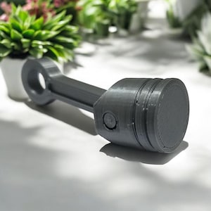 Peut inclure: Une bielle grise foncée, composant mécanique, est présentée sur une surface blanche. La tige a une ouverture circulaire et une tête cylindrique avec des rainures. Il s'agit probablement d'un objet décoratif ou d'un modèle.