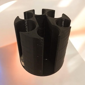 Puede incluir: Un portavasos cilíndrico negro con seis compartimentos circulares. El soporte está hecho de un material sólido y mate y tiene un diseño abierto único. Los compartimentos están espaciados uniformemente alrededor del eje central, proporcionando una solución de almacenamiento práctica y elegante.