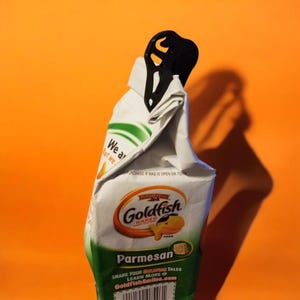 May include: A black plastic bag clip is attached to a bag of Goldfish Baked Parmesan crackers. The bag is white with green and orange accents. The text on the bag reads "Goldfish Baked Parmesan" and "Share your Goldfish Tales Learn More GoldfishSmiles.com".