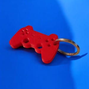 Puede incluir: Un llavero rojo con forma de mando de videojuegos. El mando tiene varios botones y un agujero circular. El llavero está unido a un anillo dorado. El fondo es azul liso.