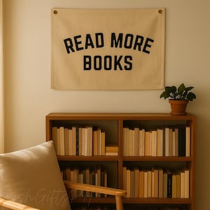 Puede incluir: Una pancarta beige con las palabras "READ MORE BOOKS" en letras negras cuelga sobre una estantería de madera llena de libros. Una pequeña planta en maceta se encuentra encima de la estantería. La escena evoca un rincón de lectura acogedor.