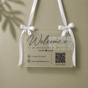 May include: Clear acrylic sign with the word "Welcome" in elegant script, "BY APPOINTMENT ONLY", a phone number, website, and a QR code. White ribbon is attached at the top with bows for hanging.