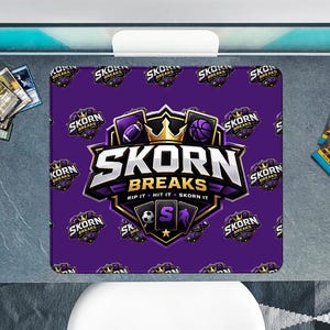 May include: A purple mousepad with the text "SKORN BREAKS" and various sports-related icons. The mousepad is surrounded by trading cards and two packs of cards, all on a gray surface.