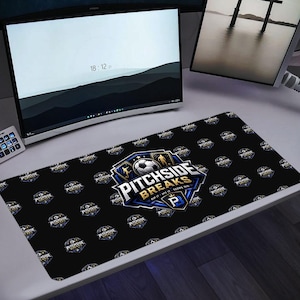 May include: Black rectangular desk mat with a repeating pattern of a soccer-themed logo. The central logo features a soccer ball and the text "PITCHSIDE BREAKS" in white and blue. A computer monitor and other tech accessories are in the background.