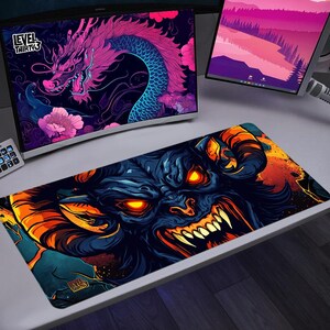 May include: A rectangular gaming mousepad with a detailed illustration of a demonic creature in black and blue, with orange glowing eyes and sharp teeth. The mousepad is on a white desk, with computer monitors in the background.