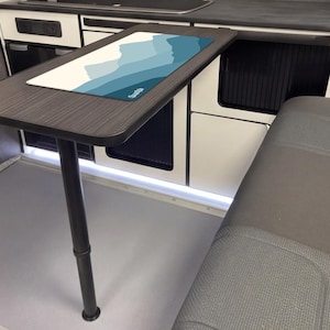 May include: A rectangular table with a dark wood grain finish and a single black support leg. The table has a rectangular mat with a mountain range design in shades of blue and white. The table is in a camper van.