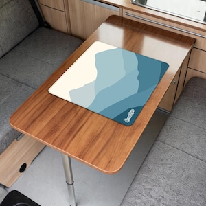 May include: A rectangular mousepad featuring a blue and cream mountain range design. The mousepad rests on a wooden table with a silver leg. A small white logo is in the bottom right corner of the mousepad.