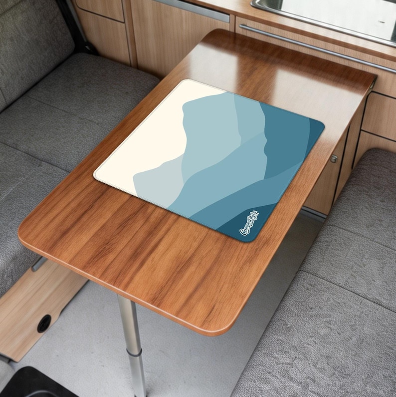 May include: A rectangular mousepad featuring a blue and cream mountain range design. The mousepad rests on a wooden table with a silver leg. A small white logo is in the bottom right corner of the mousepad.