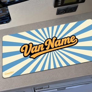 May include: A rectangular desk mat featuring a light blue and cream-coloured sunburst pattern. The text "Van Name" is printed in a bold, orange, cursive font across the centre. The small logo "CamperStylz" appears in the bottom left corner.