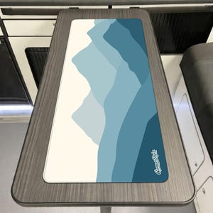 May include: A rectangular desk mat with a mountain range design in shades of blue and white. The mat has a white border and the words "CamperStylz" in white script in the lower right corner. The mat is placed on a wooden table.