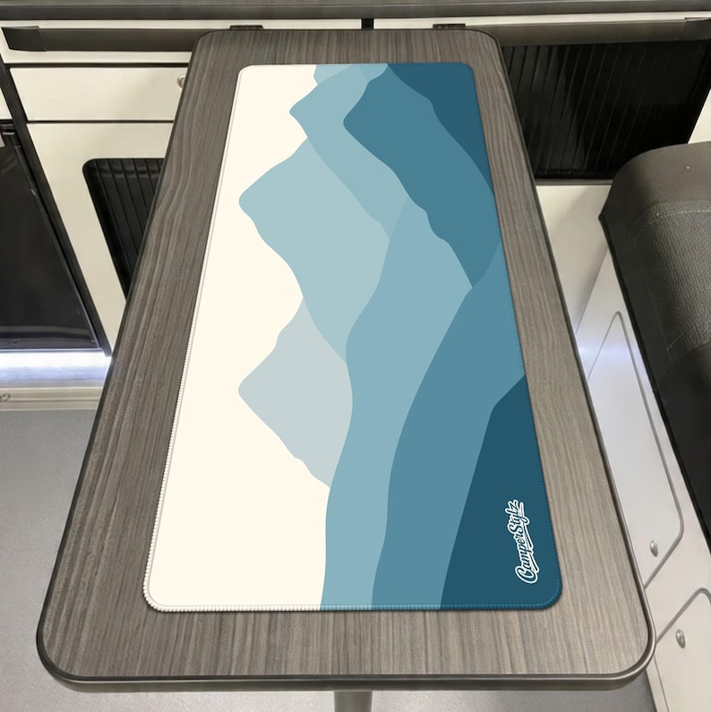 May include: A rectangular desk mat with a mountain range design in shades of blue and white. The mat has a white border and the words "CamperStylz" in white script in the lower right corner. The mat is placed on a wooden table.