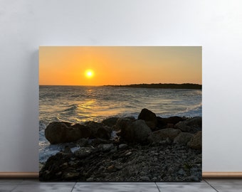 Könnte beinhalten: Ein malerischer Sonnenuntergang über einem felsigen Strand, wobei die Sonne über dem Meer untergeht. Das Wasser ist tiefblau und der Himmel leuchtend orange und gelb.