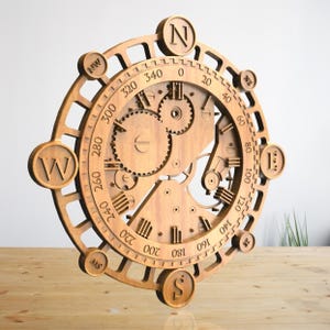 Pode incluir: Uma bússola de madeira com um mecanismo de engrenagem detalhado e uma rosa dos ventos com as direções cardeais N, S, E e W. A bússola é marcada com números de 0 a 360 graus.