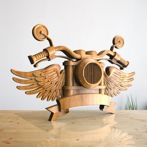 May include: Wooden motorcycle sculpture with wings and a banner. The motorcycle is detailed with a headlight and handlebars.