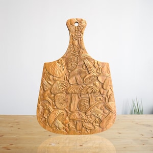 以下が含まれることがあります： キノコの彫刻が施された木製のまな板。まな板は薄い茶色で、持ち手があります。