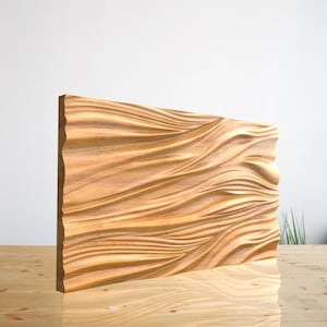 May include: A rectangular wooden wall art piece with a carved wave pattern. The artwork is made of light-colored wood and stands upright on a wooden surface. The edges of the piece have a rough, natural texture.