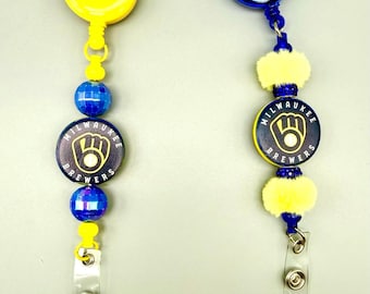 Puede incluir: Dos portatarjetas retráctiles con logotipos de los Milwaukee Brewers. El de la izquierda tiene carcasa amarilla, cuentas azules facetadas y un dije con logo. El de la derecha tiene carcasa azul, pompones amarillos, cuentas azules y un dije.