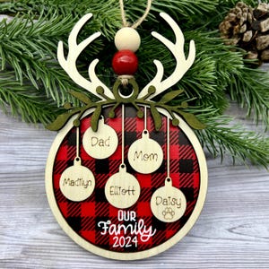 May include: Wooden Christmas ornament with a red and black buffalo plaid pattern. The ornament features the words "Our Family 2024" and personalized name tags. It is adorned with wooden antlers, green leaves, and red and beige beads.