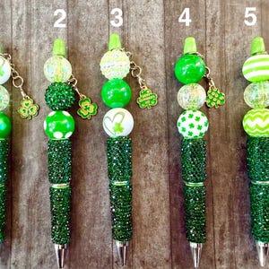 May include: Five decorative pens with green and white beads and shamrock charms. The pens have a green, jewel-encrusted barrel and a silver tip. Each pen has a different design, including polka dots, stripes, and heart-shaped shamrocks.