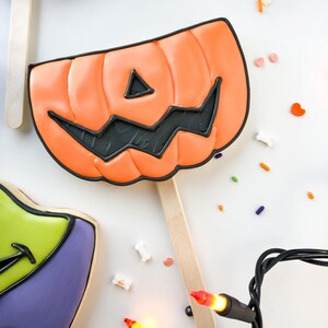 Op de afbeelding: Oranje pompoenvormige koek op een houten stok, versierd met zwart glazuur voor het gezicht. De koek is omgeven door kleurrijke hagelslag en een slinger van Halloween-lichtjes. Andere koekjes zijn zichtbaar in de afbeelding.