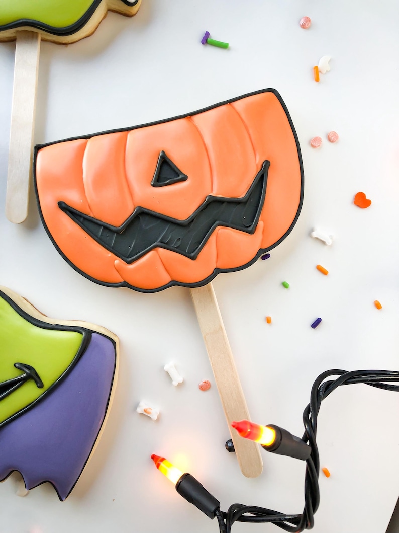Op de afbeelding: Oranje pompoenvormige koek op een houten stok, versierd met zwart glazuur voor het gezicht. De koek is omgeven door kleurrijke hagelslag en een slinger van Halloween-lichtjes. Andere koekjes zijn zichtbaar in de afbeelding.